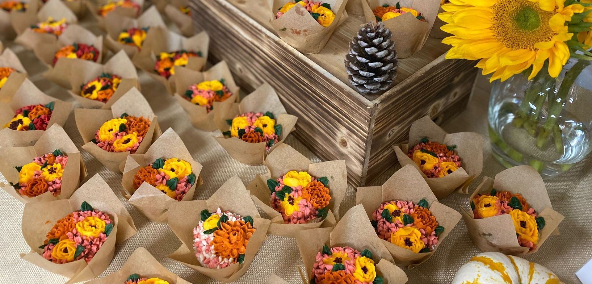 Mini cupcakes decorated with buttercream flowers in brown paper wrappers, with sunflowers and a pine cone.