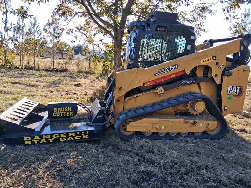 A yellow Caterpillar skid steer loader with a black brush cutter attachment sits in a field on a sunny day.