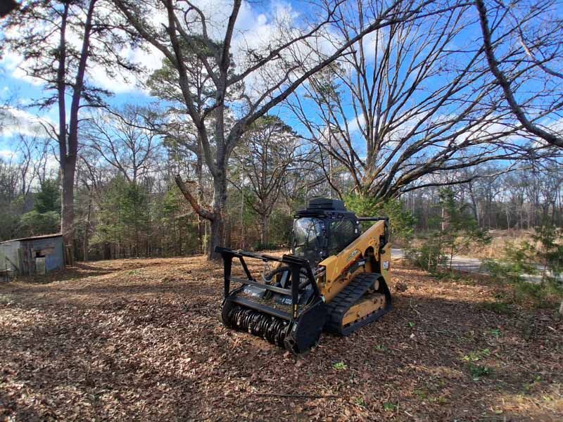A yellow track loader with a forestry mulching attachment parked in a wooded area covered in fallen leaves.