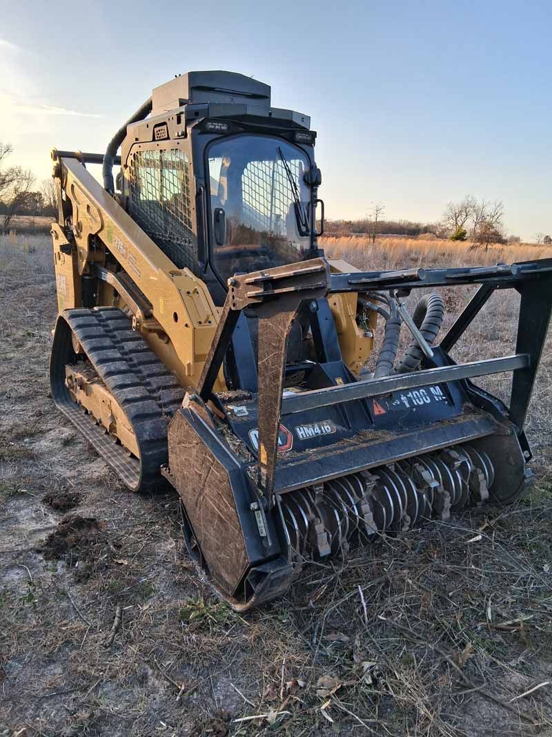 A yellow tracked skid steer loader with a heavy-duty brush cutter attachment parked in a field at sunset.