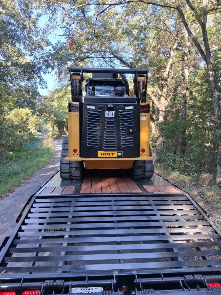 A yellow Caterpillar skid steer loader parked on a flatbed trailer on a wooded road.