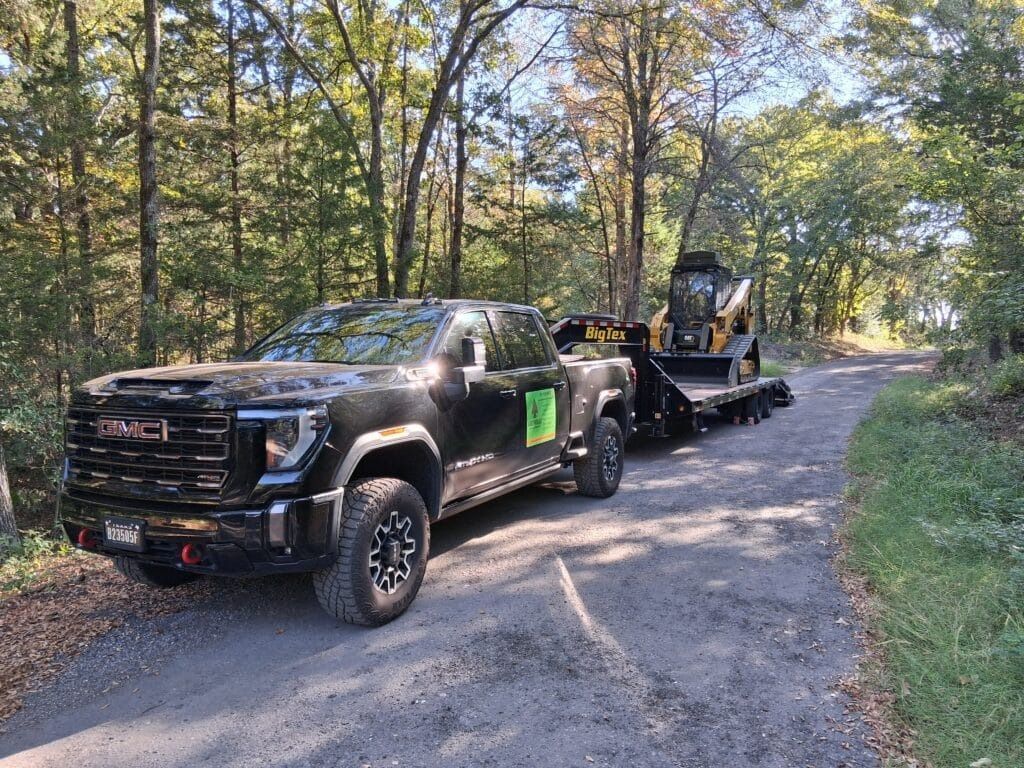 A black GMC Sierra pickup truck towing a flatbed trailer with a yellow skid steer loader on a wooded gravel road.