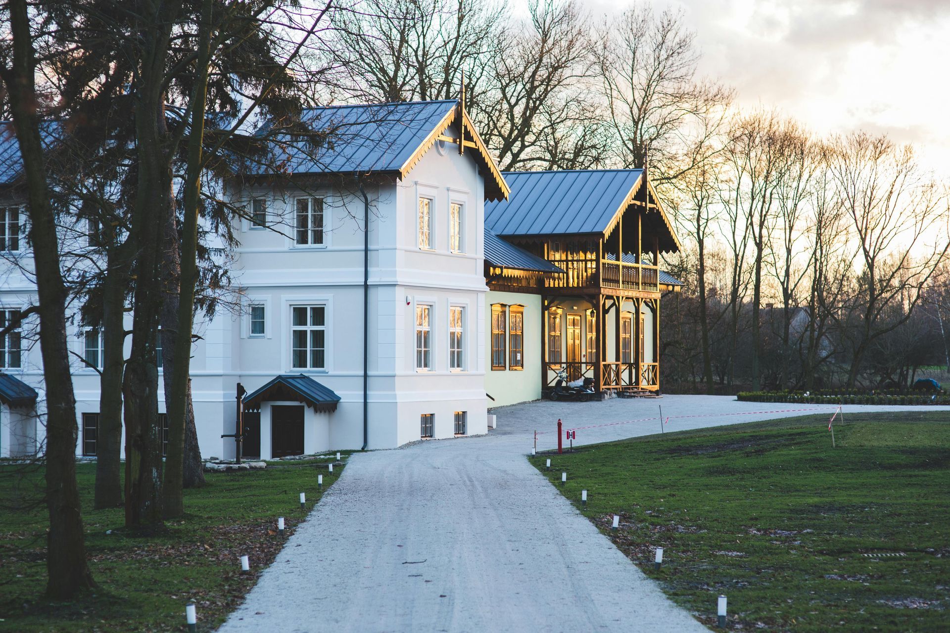 A two-story white building with a blue metal roof, an attached wooden veranda, and a gravel path leading to the entrance.