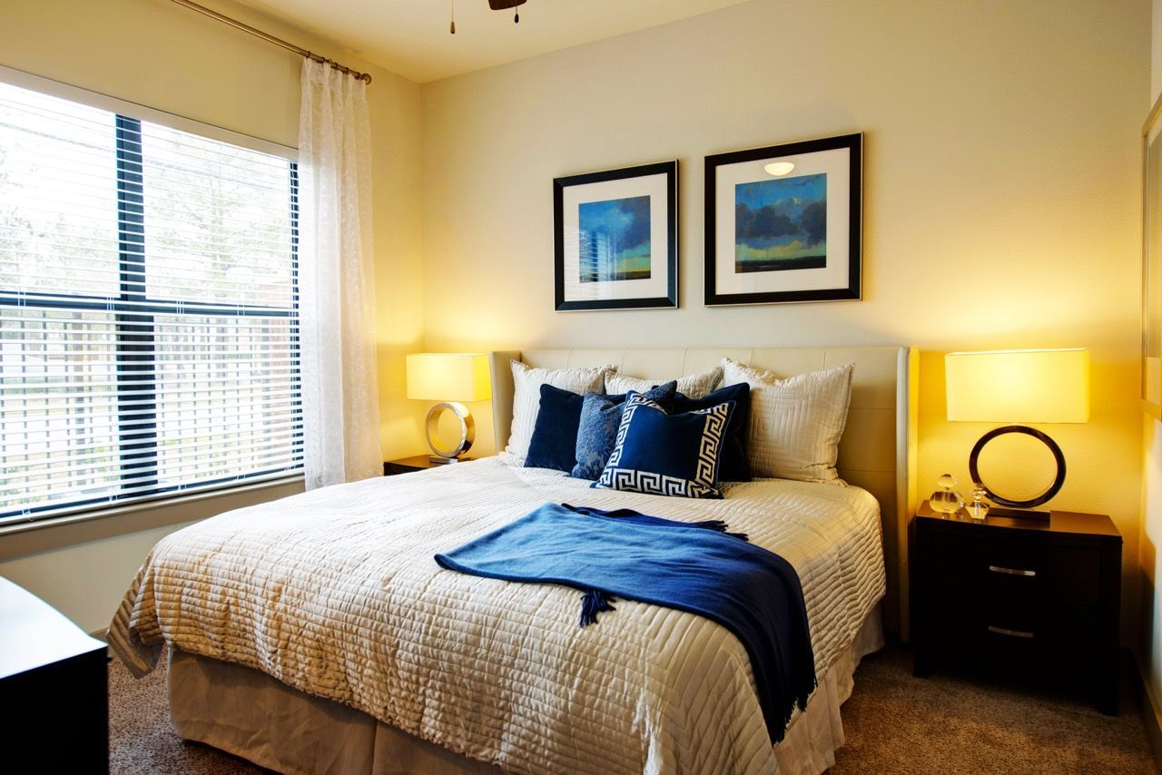 Bright bedroom with a queen bed, two nightstands, lamps, and a large window with blinds.