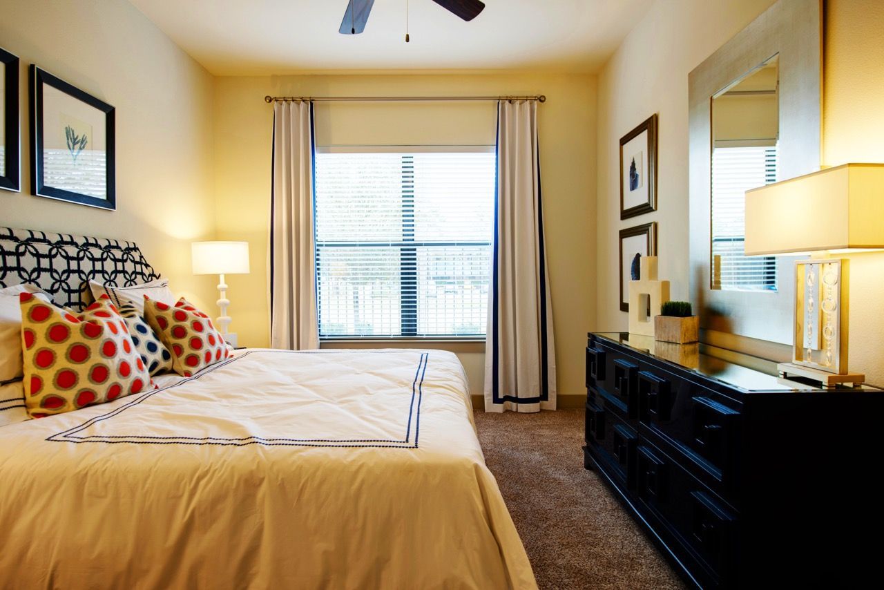 Bright bedroom in a modern apartment with a large window, bed, dresser, and decorative pillows.
