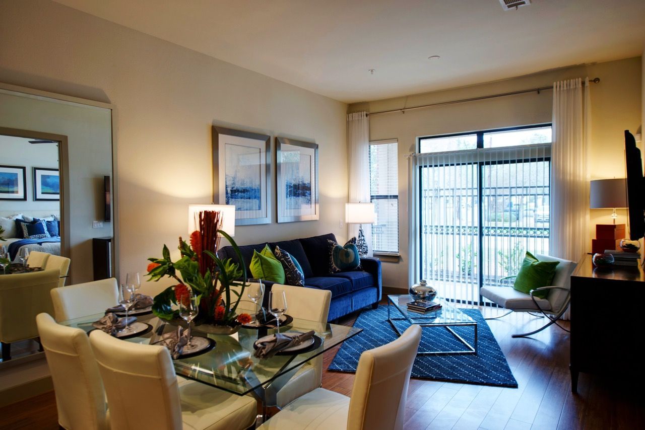 Open-concept apartment living area with glass dining table, white chairs, blue sofa, and balcony doors.