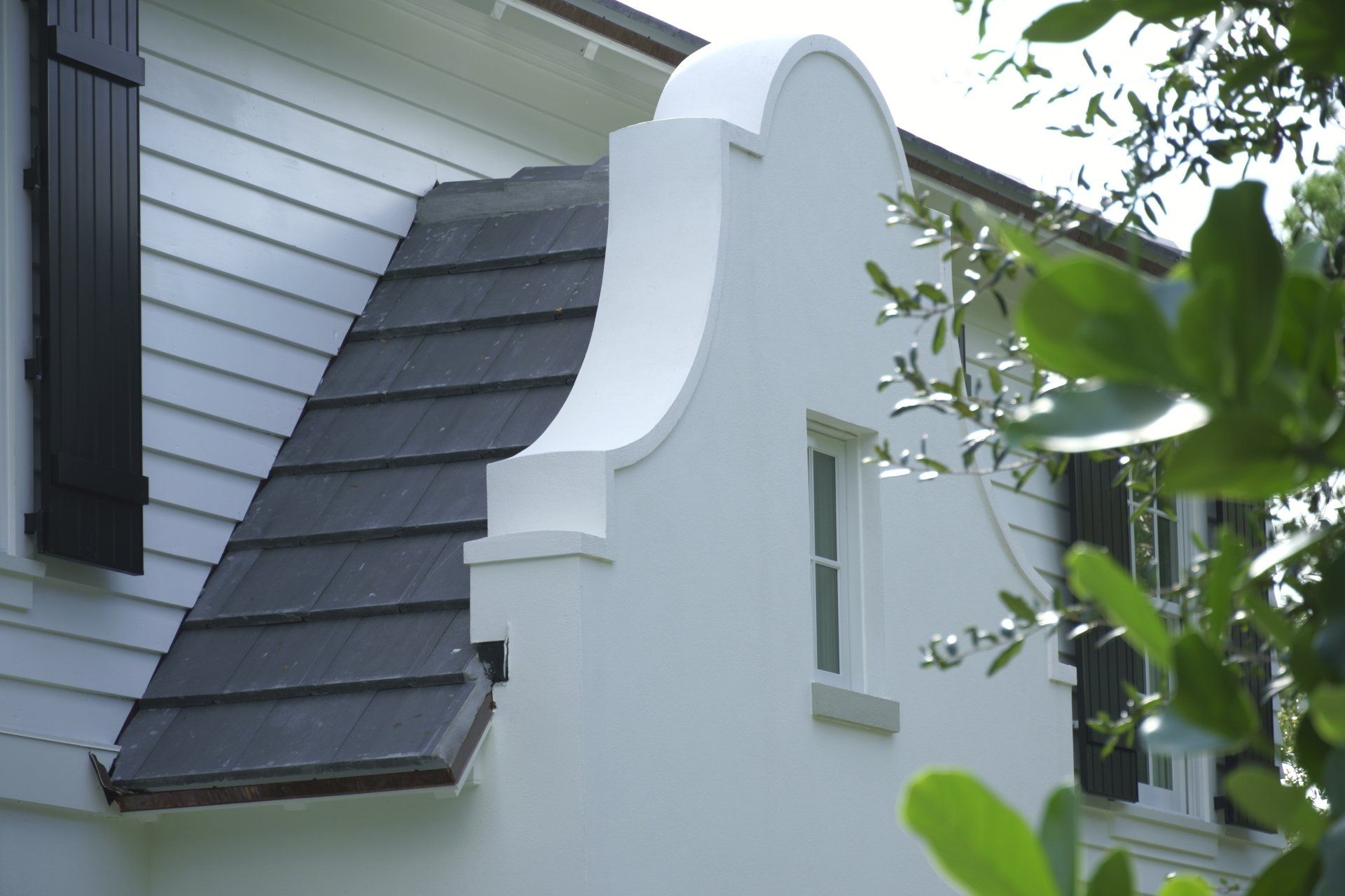 White house gable with curved facade and dark roof tiles, with a window and shutters.