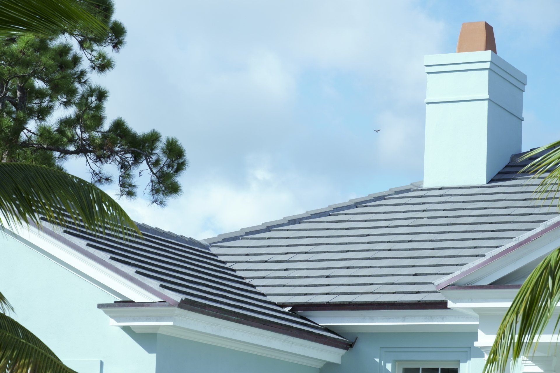 Blue house roof with gray tiles, chimney, and palm trees against a cloudy sky.