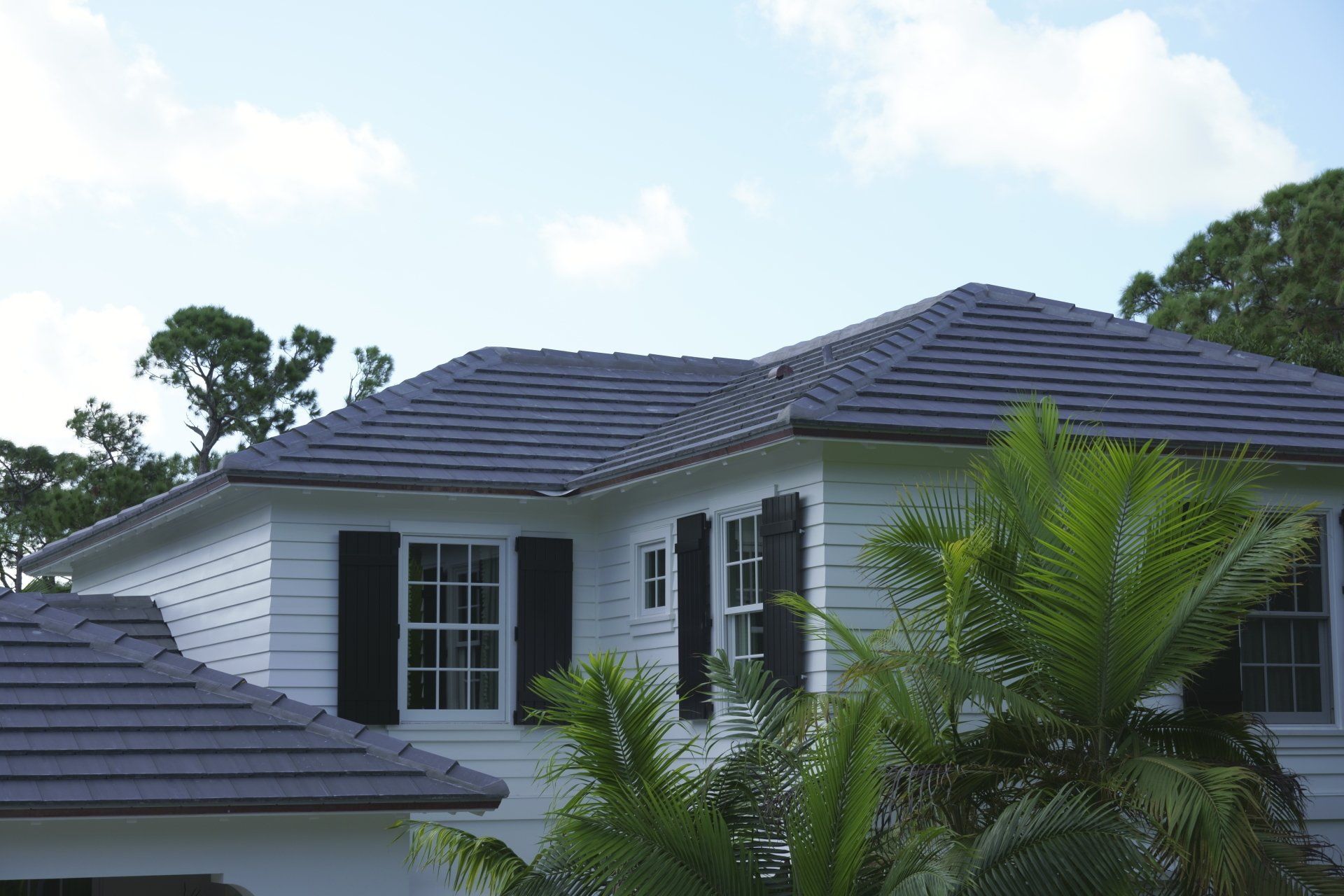 White house with gray tile roof, black shutters, and palm trees against a cloudy sky.