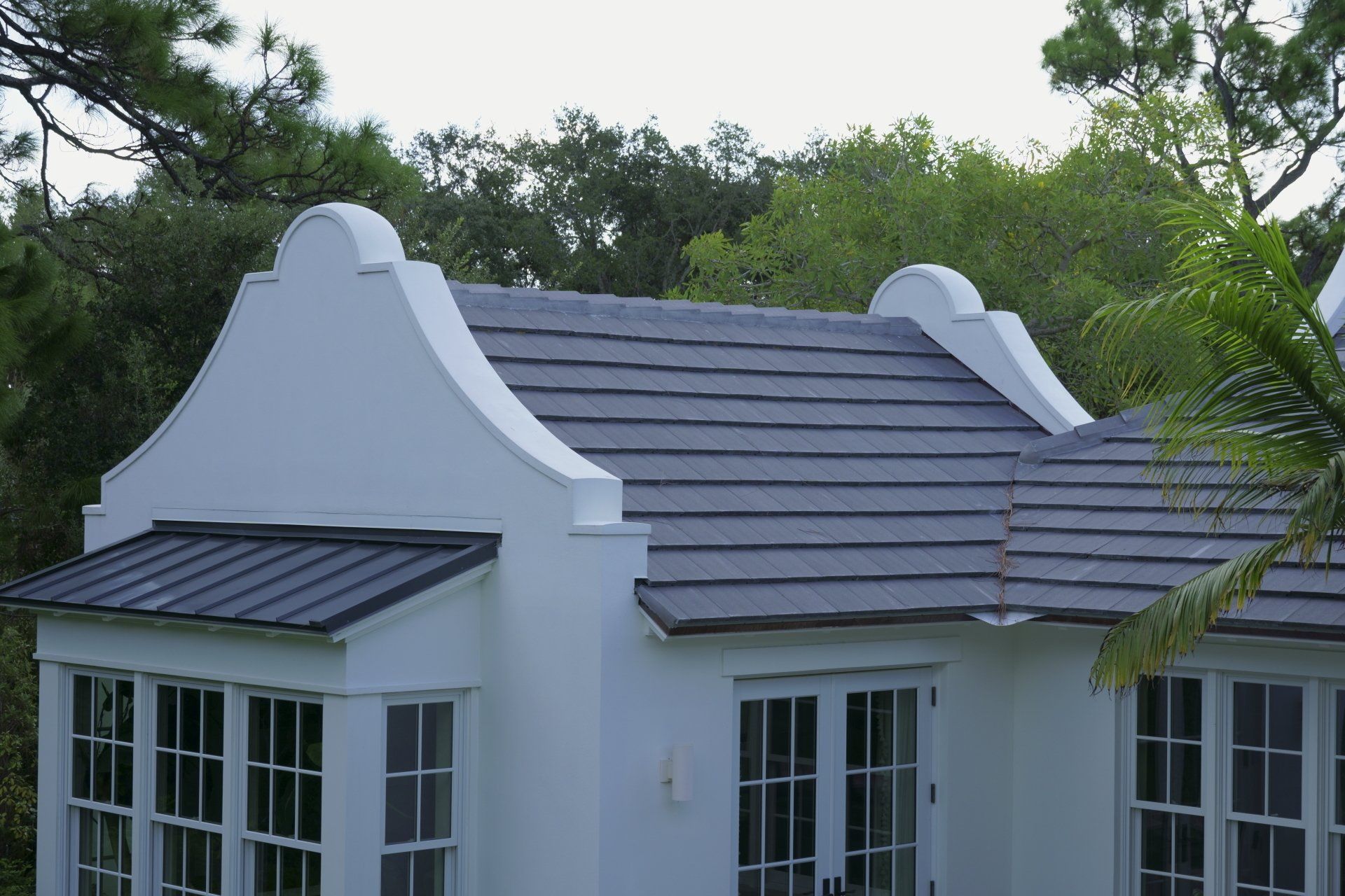 White house with gray tile roof, bay window, and arched rooftop features; surrounded by green trees.