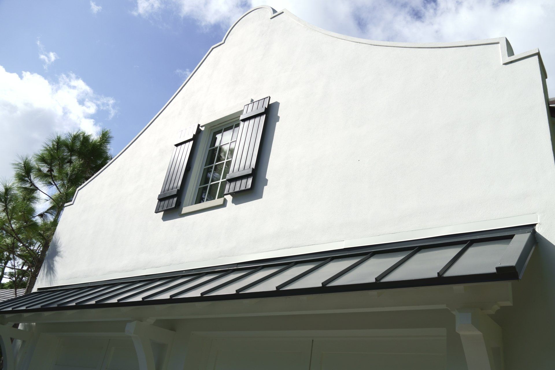 White stucco building with a black metal awning and shuttered window against a blue sky.