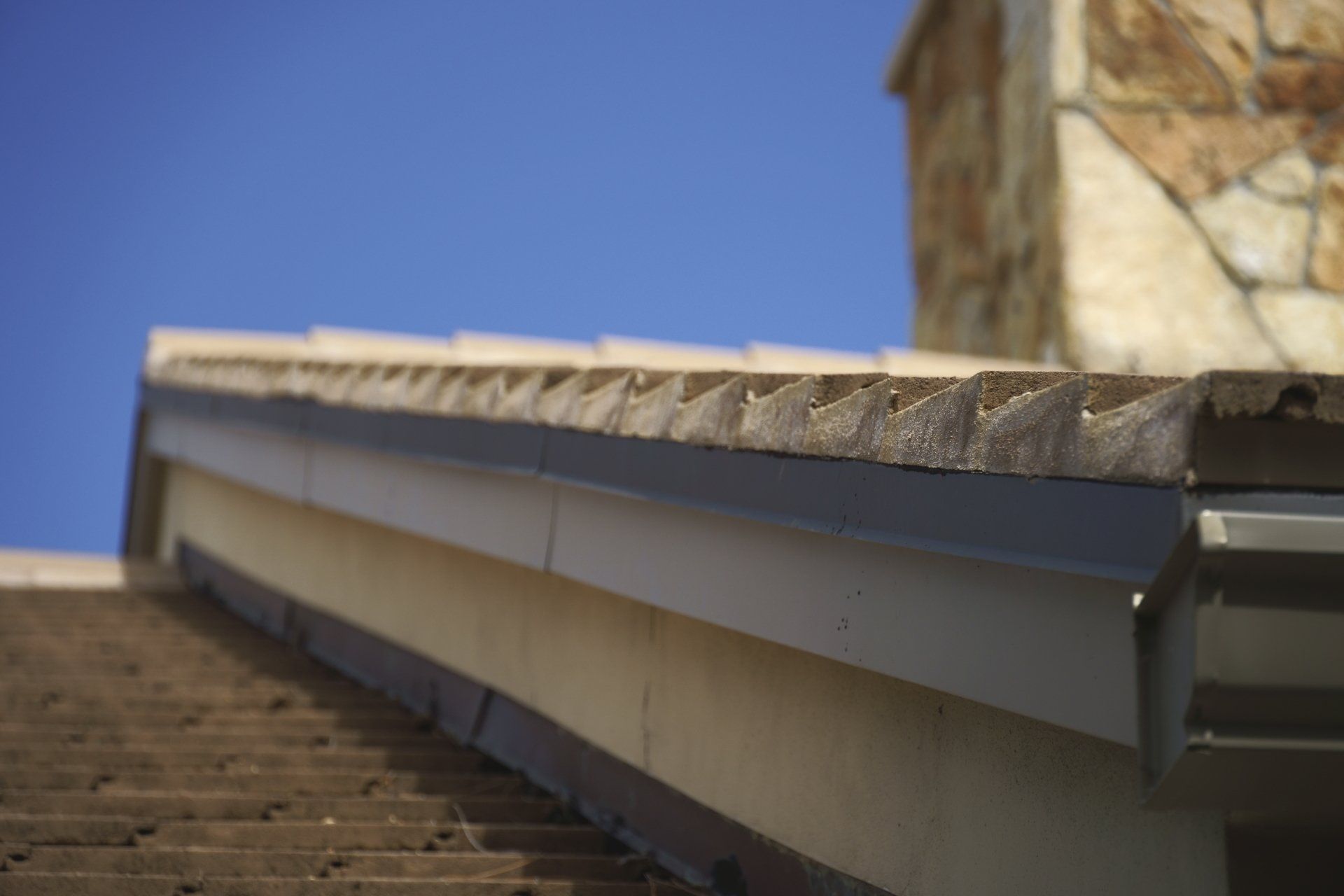 Close-up of a roof's edge with a gutter and a stone chimney against a blue sky.