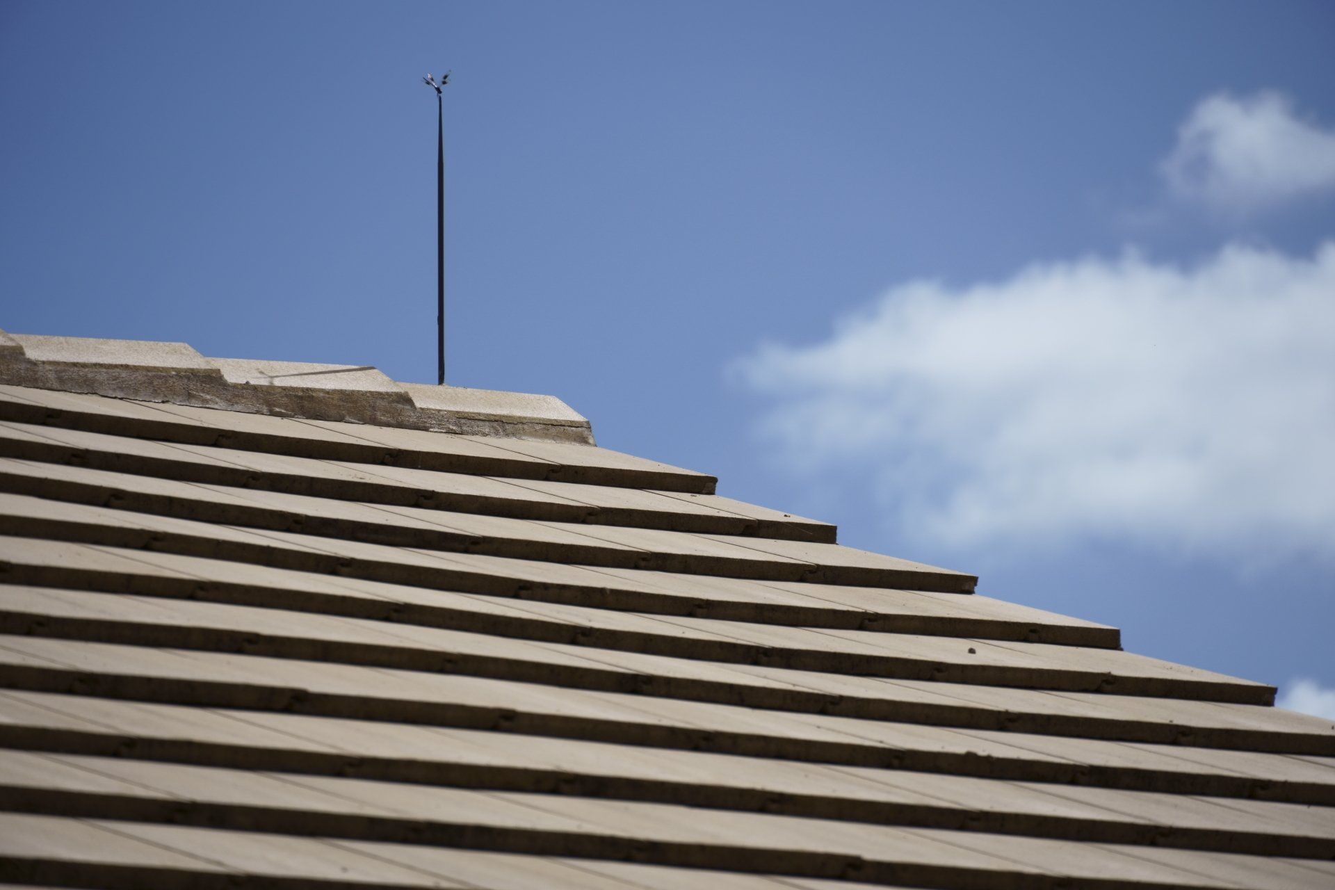 Tan tiled roof with lightning rod against a blue sky with white clouds.