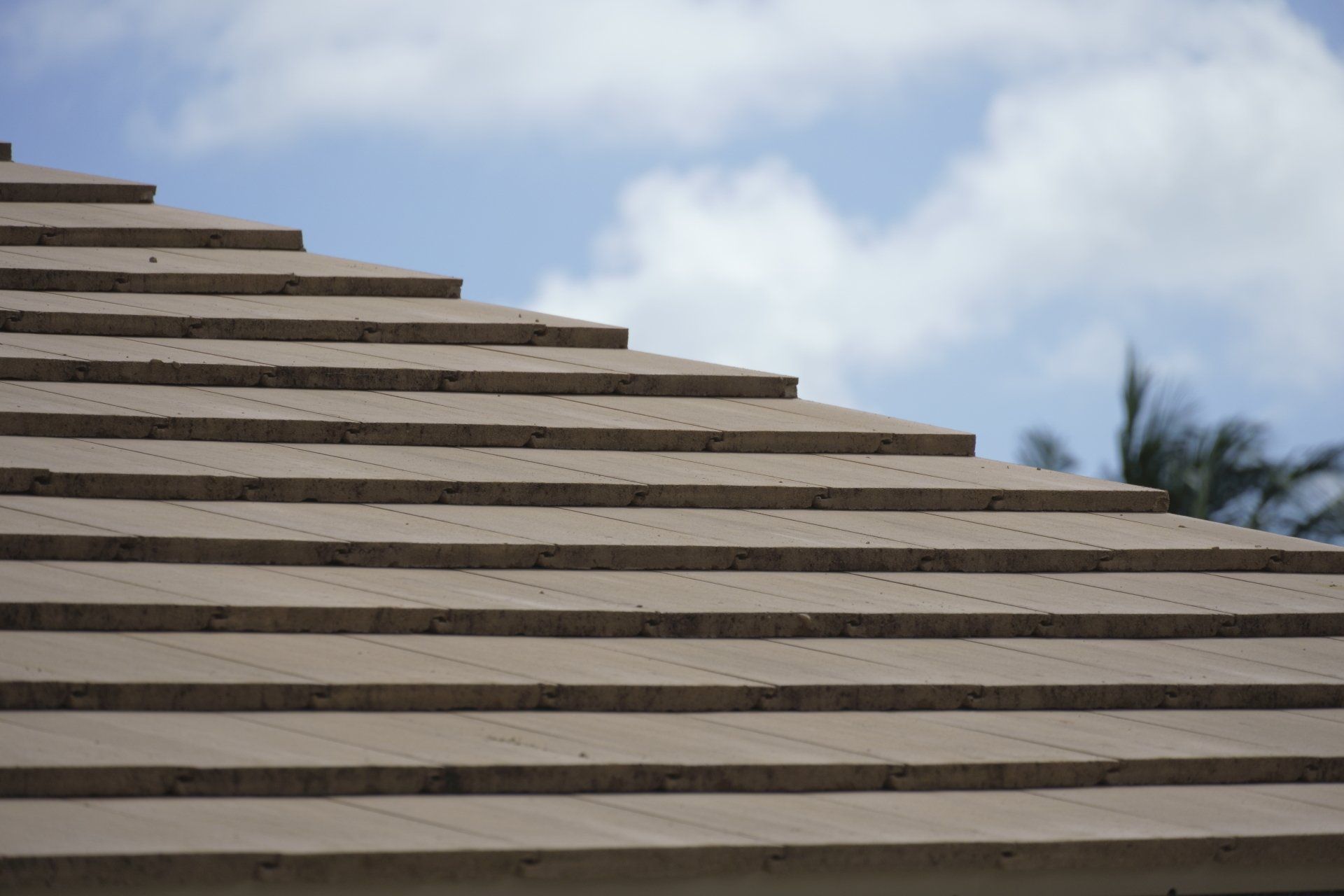 Close-up of a layered, light brown roof against a blue sky with fluffy clouds.