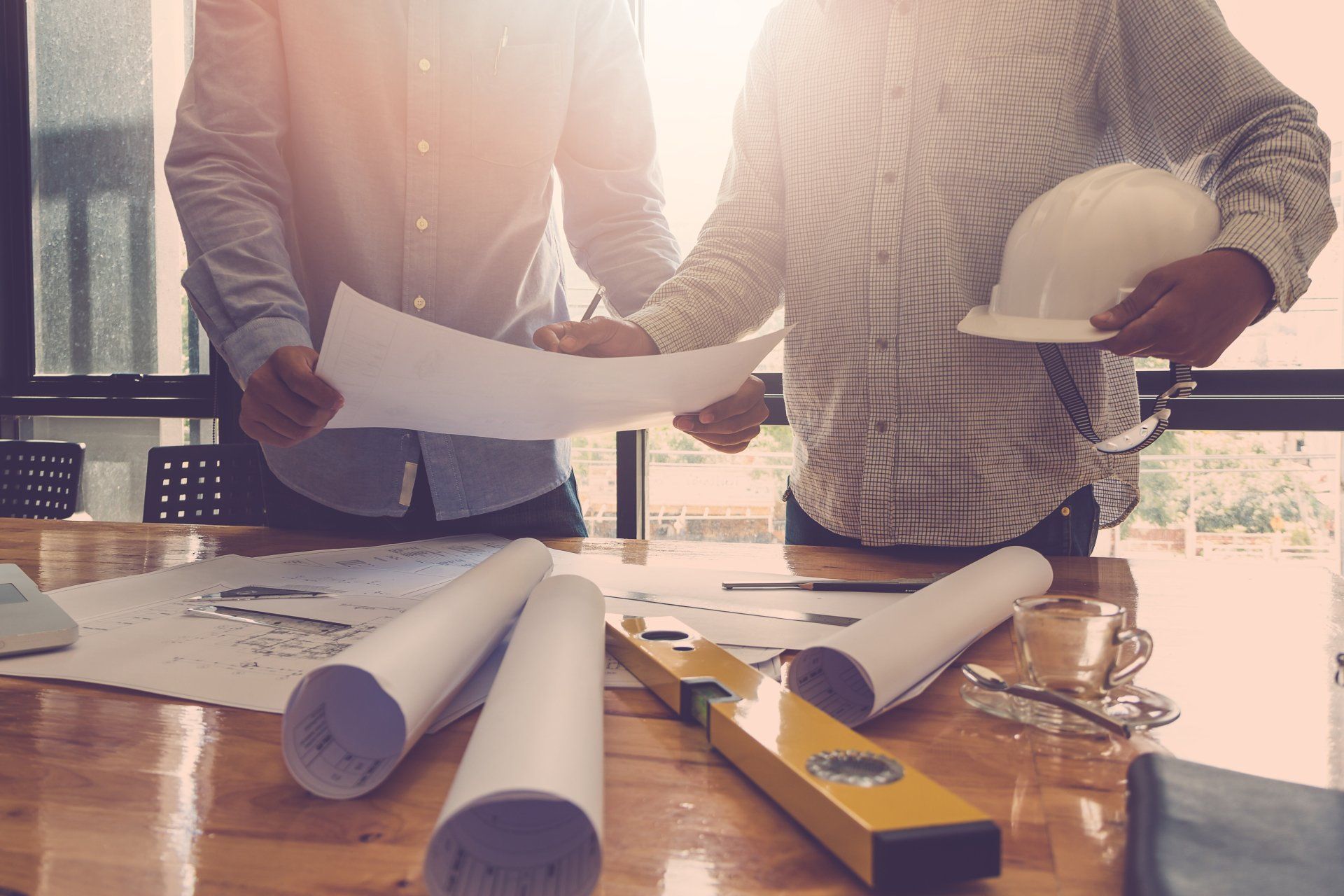 Two people reviewing blueprints at a table, construction plans and tools visible. Sunlight streams in.