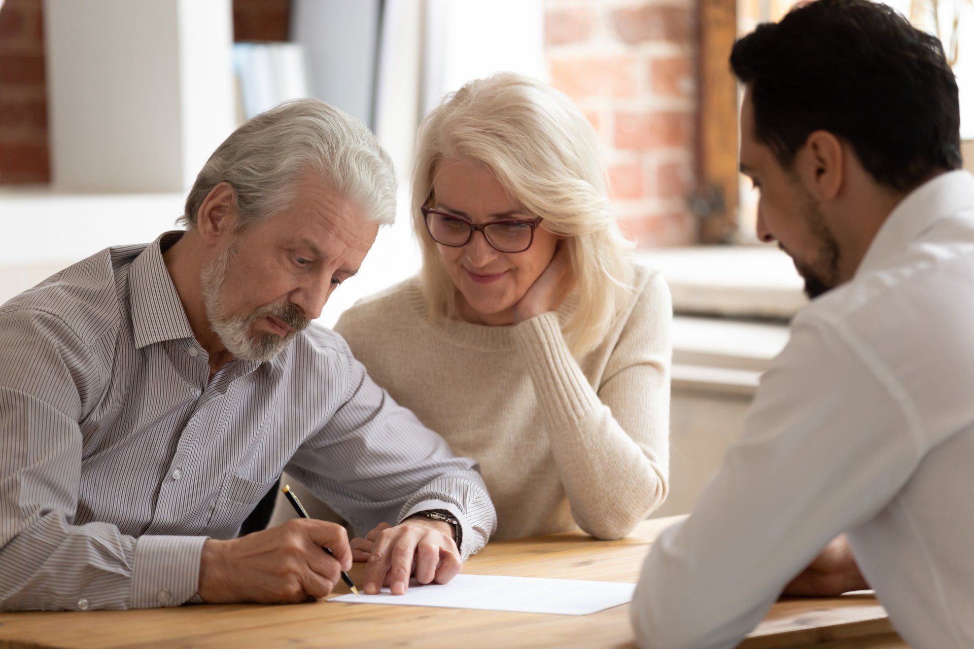 Senior couple signing document at a table with a man in a light-colored shirt. Brick wall in the background.