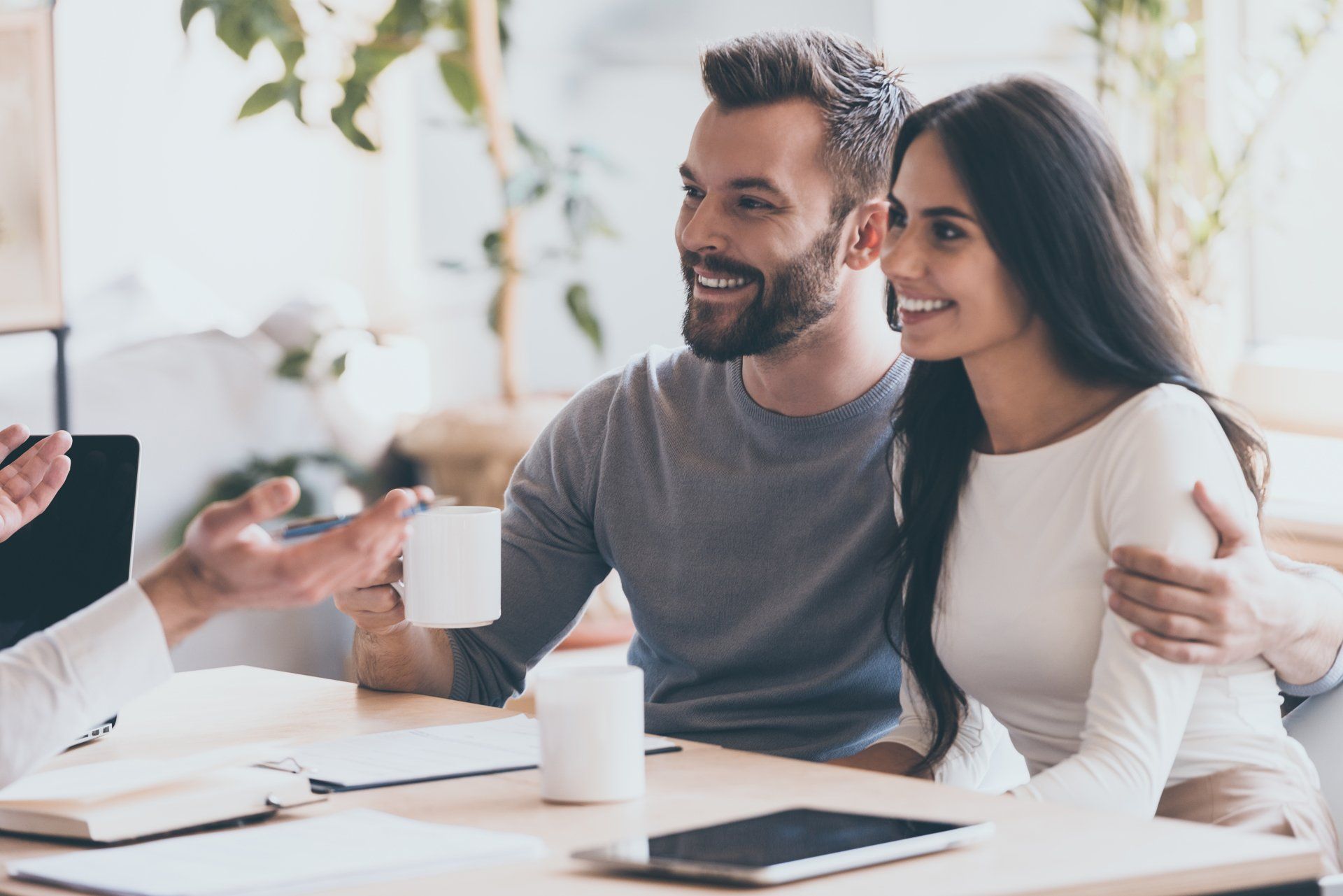 Couple smiling, arms around each other, at a table with a person gesturing with a cup; sunny room.