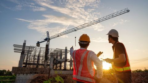 Two people in hard hats and safety vests stand at a construction site at sunset, discussing plans before a crane.