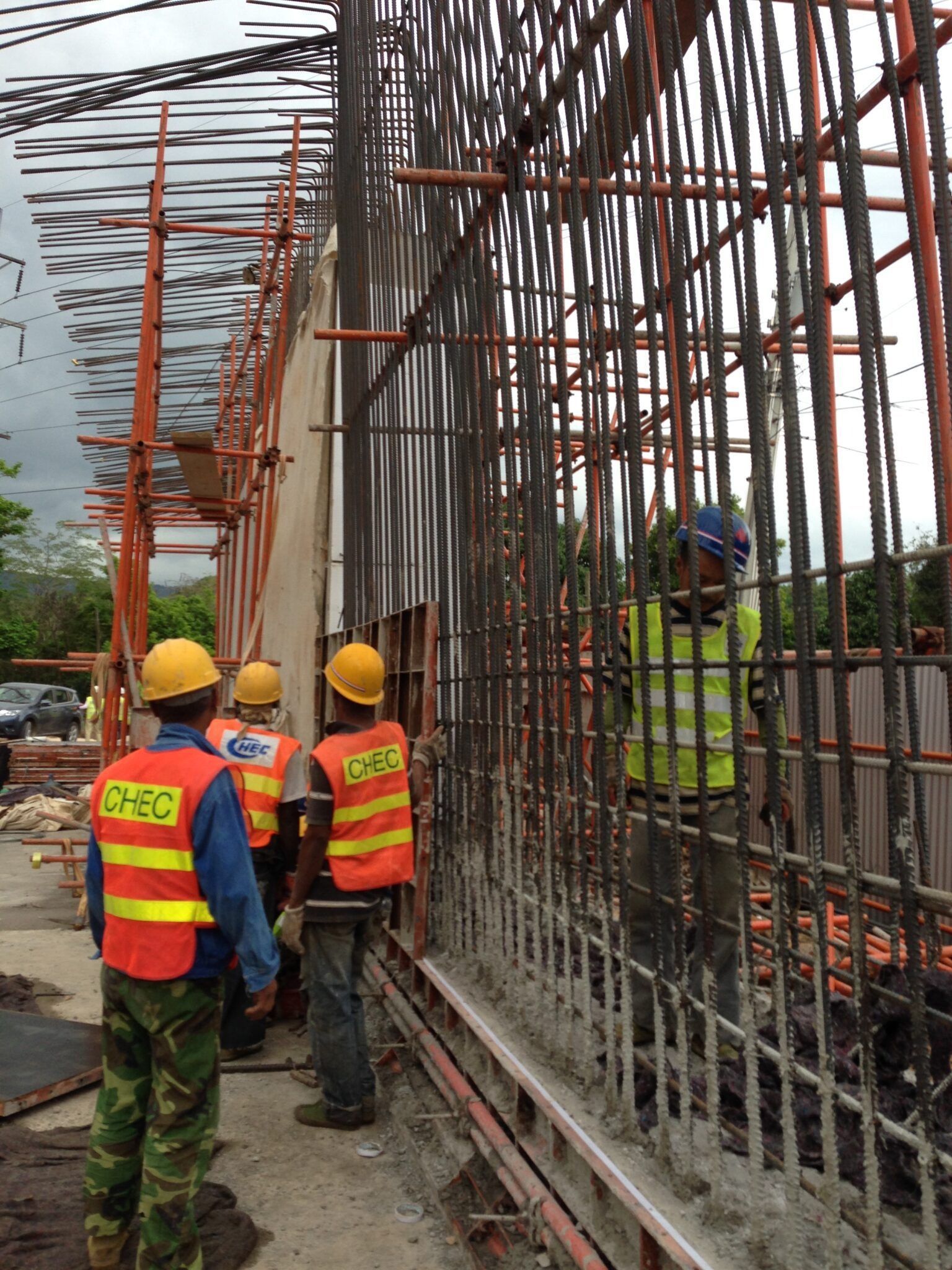 Construction workers in high-visibility safety gear assemble vertical steel rebar reinforcement for a wall on a work site.
