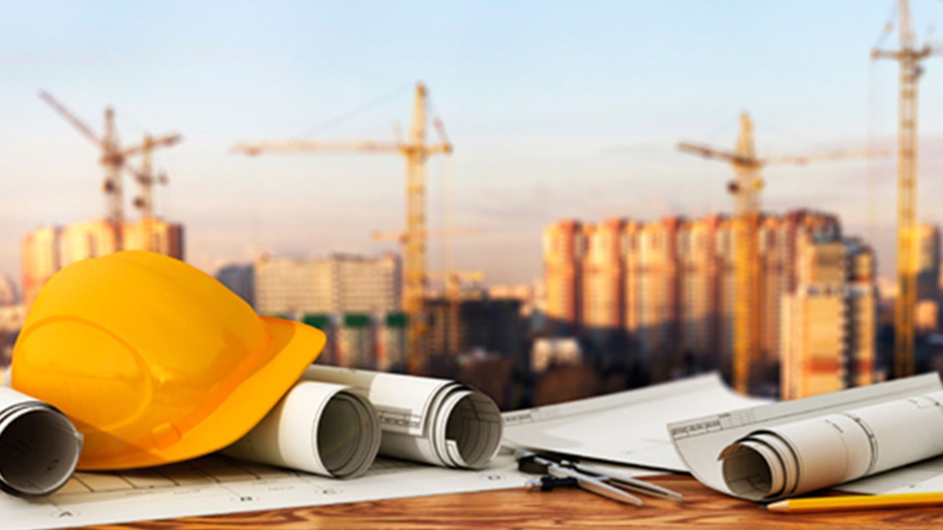A yellow hard hat and rolled-up architectural blueprints sit on a wooden table before a blurred construction site.