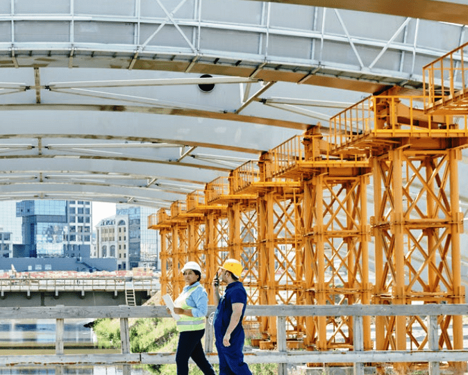 Two construction workers in safety gear walk on a bridge site near yellow support structures and a city skyline.