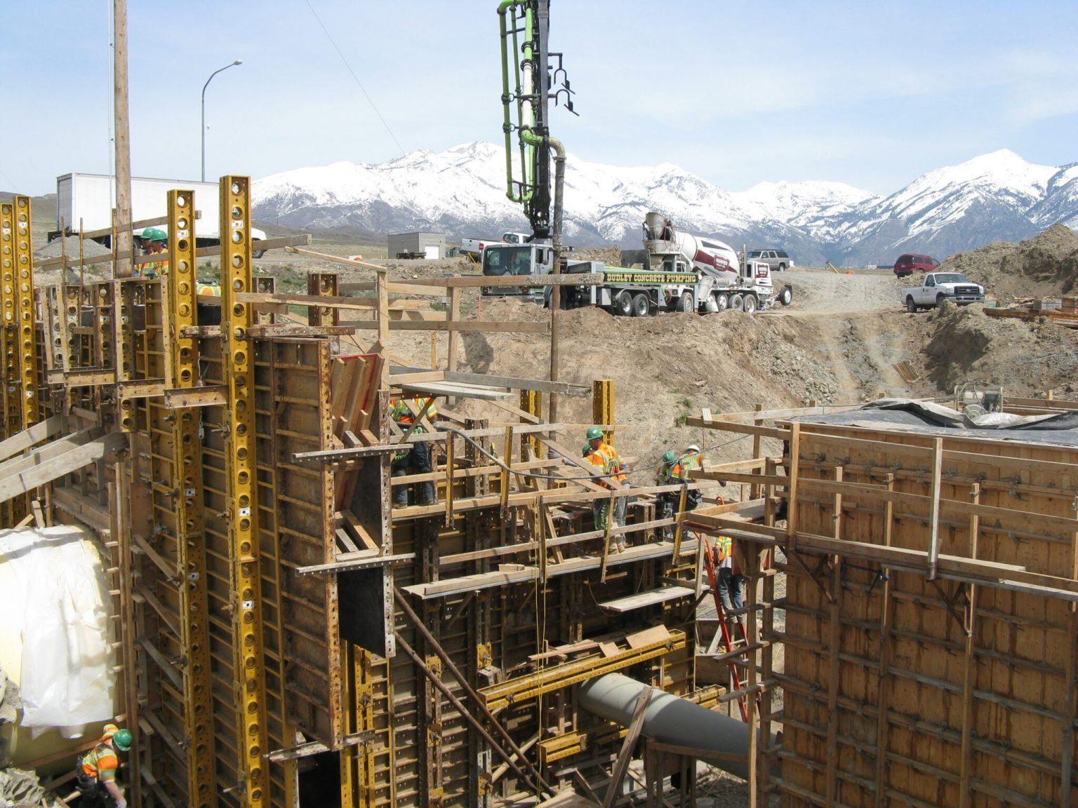 Construction crew working on concrete formwork for a wall at a site with a concrete pump and snow-capped mountains.