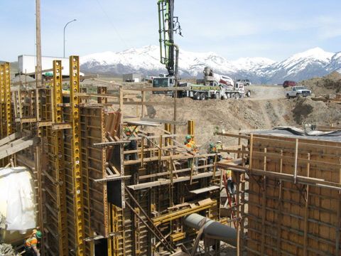Construction site with workers, tall metal formwork, and a drill rig against a backdrop of snow-capped mountains.