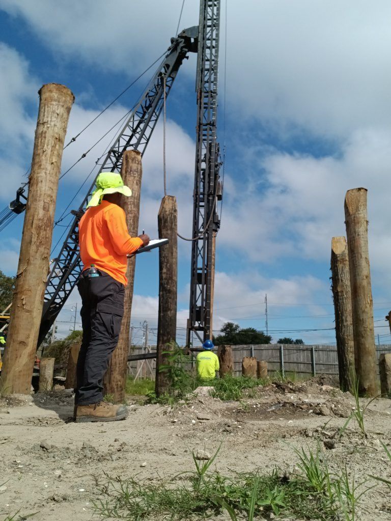 A construction worker in a high-visibility shirt observes a crane driving wooden piles into the ground at a site.