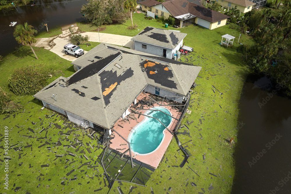 An aerial view of a house with significant roof damage and debris scattered across the lawn following a storm.