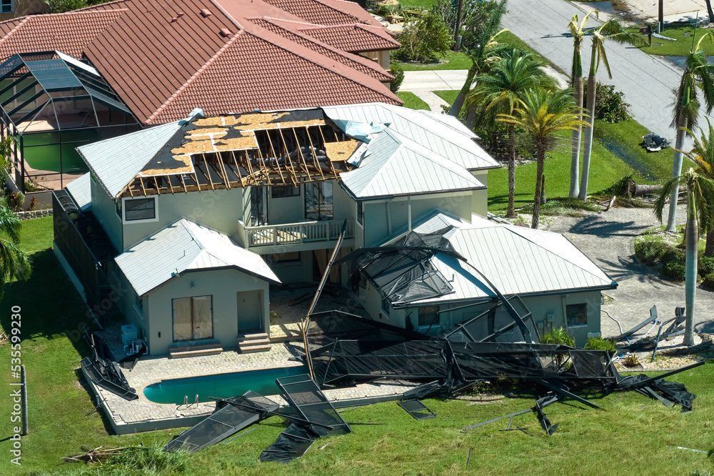 Aerial view of a hurricane-damaged home with missing roof tiles, exposed rafters, and a collapsed pool screen enclosure.