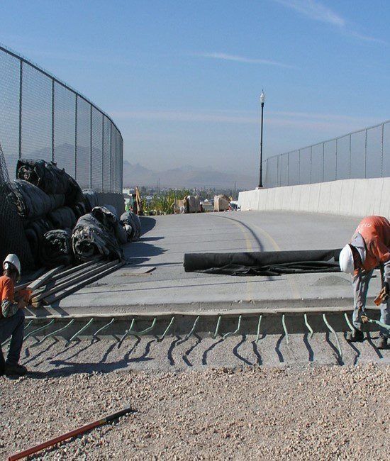 Two construction workers work on a road bridge expansion joint, with rebar exposed and piles of materials nearby.