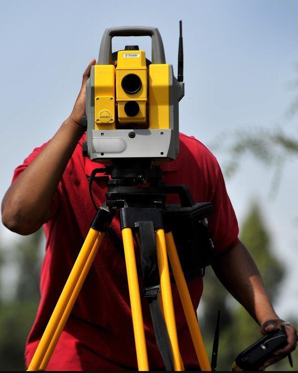 A person in a red shirt adjusts a yellow survey total station mounted on a tripod against a clear sky.