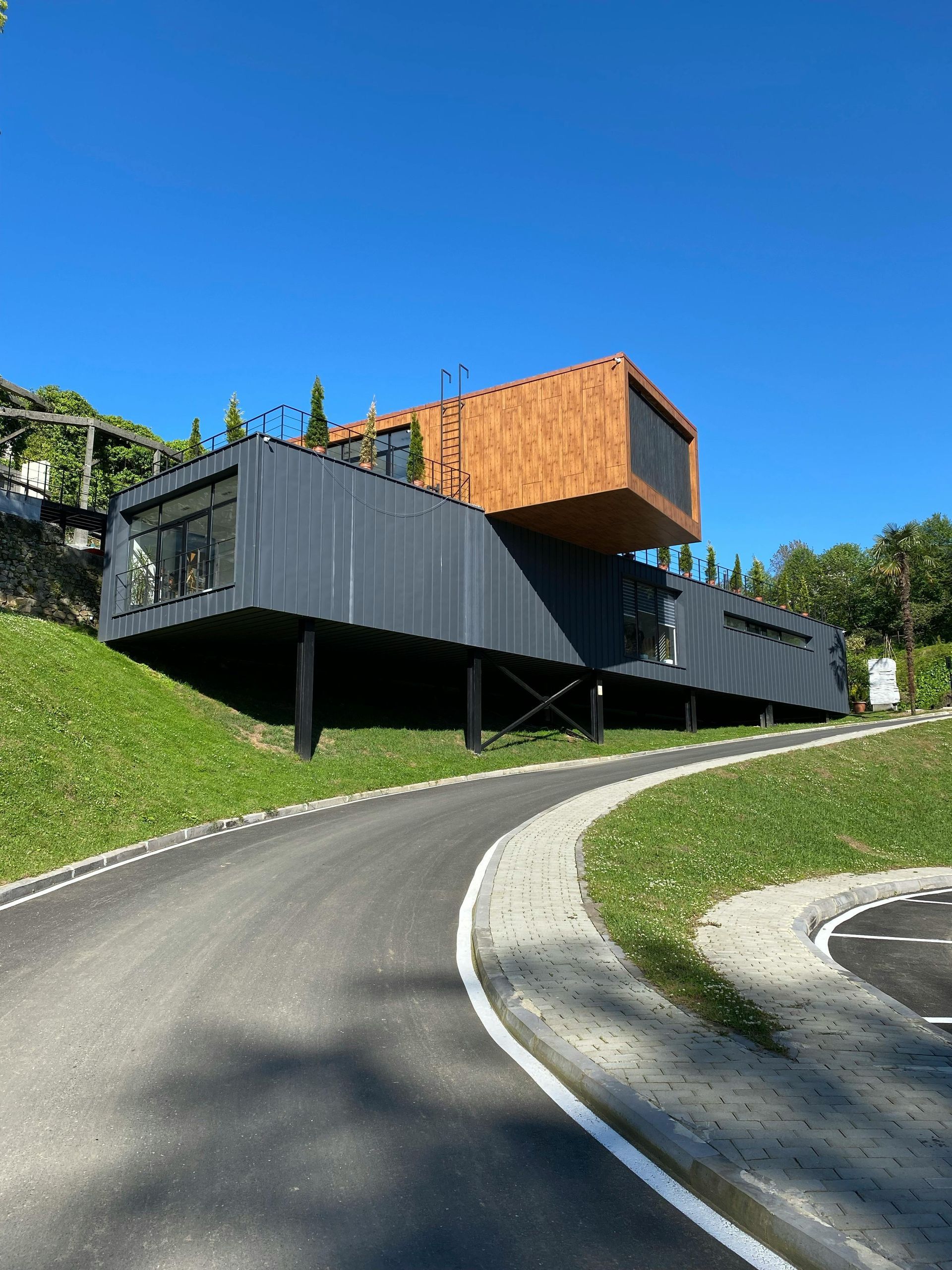 A modern house with a charcoal gray lower level and wooden top level, elevated on a grassy hill beside a paved road.