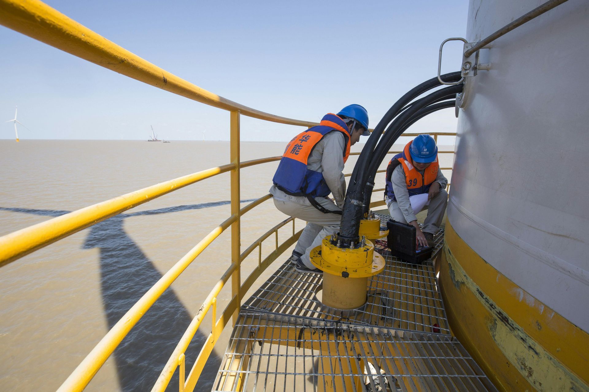 Two technicians in hard hats and life vests perform maintenance on cables on an offshore wind turbine platform.