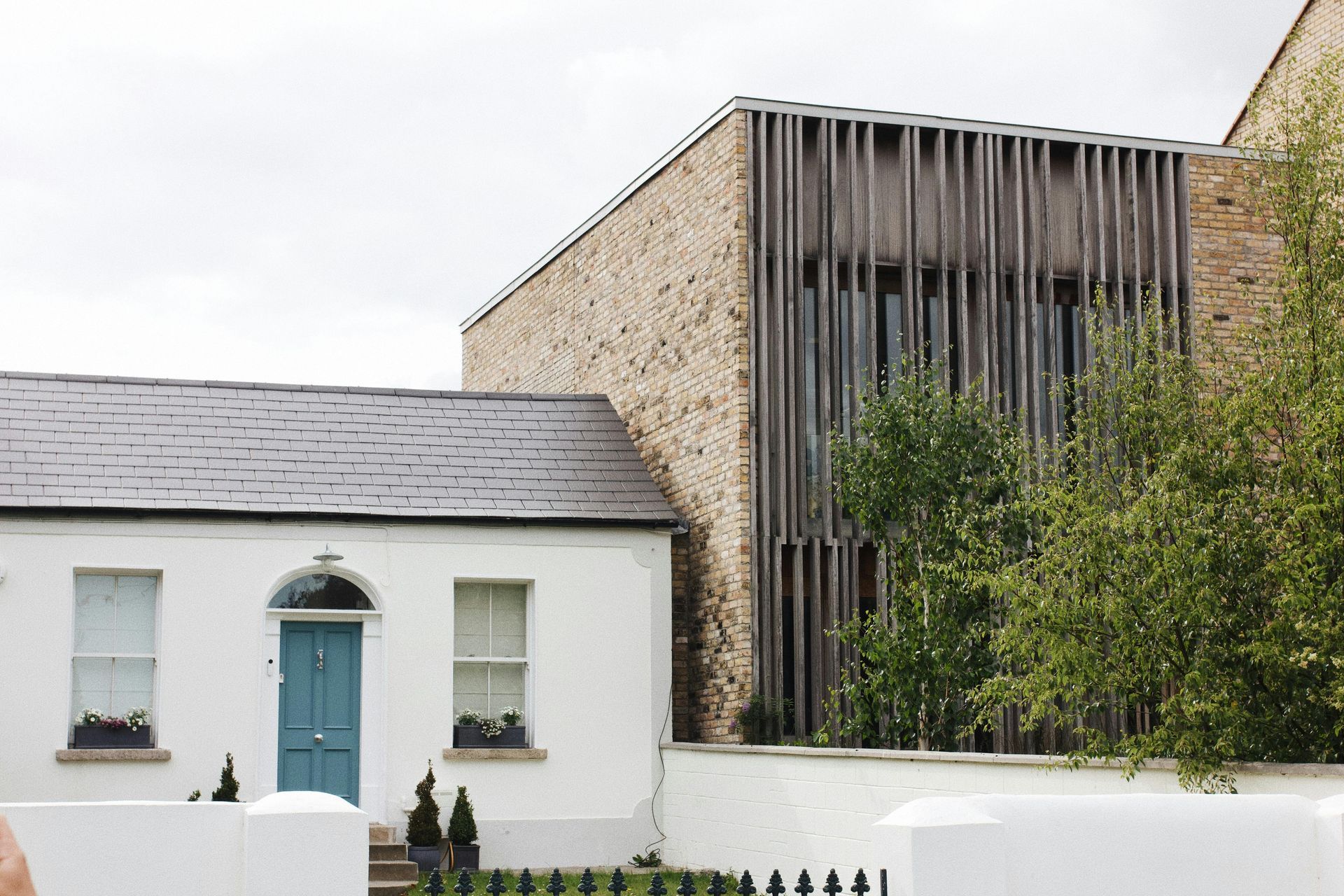 A historic, white cottage with a blue door sits next to a modern, stone-clad extension with tall, vertical wooden slats.