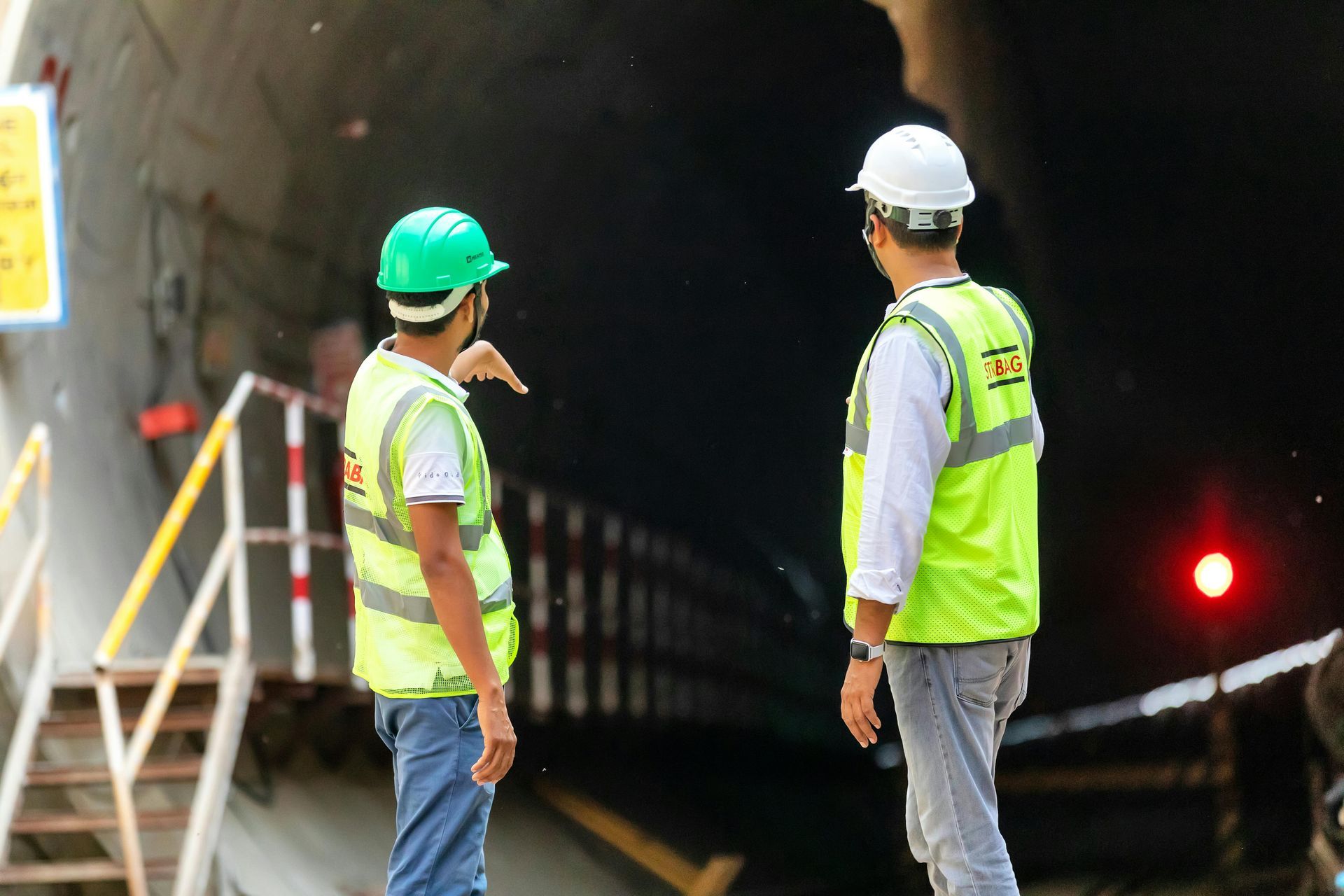 Two workers in high-visibility vests and hard hats stand outside a tunnel entrance, with one gesturing toward the tunnel.