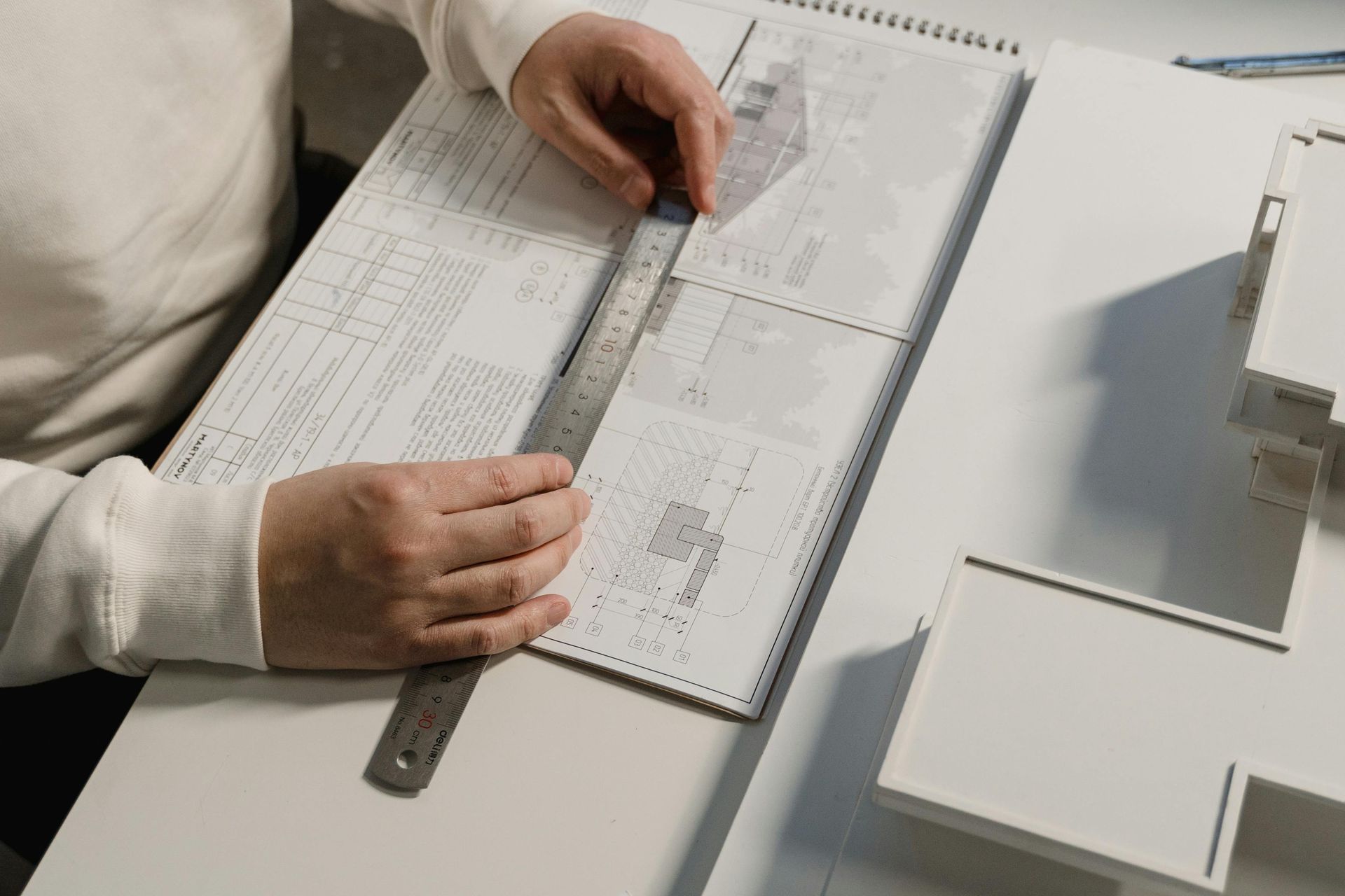A person's hands use a metal ruler to measure technical architectural drawings on a white desk next to a building model.