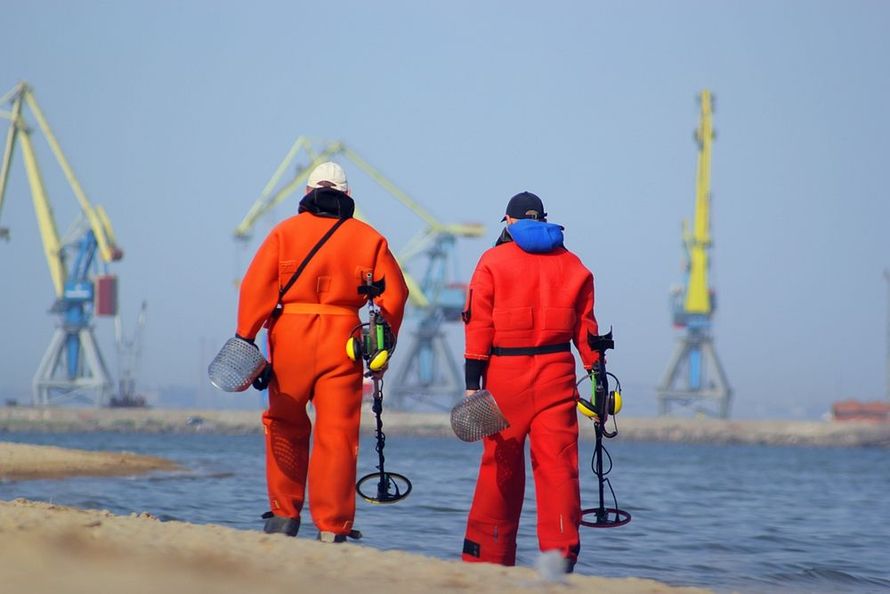 Two workers in orange safety suits walking along a shoreline with equipment and cranes in the background