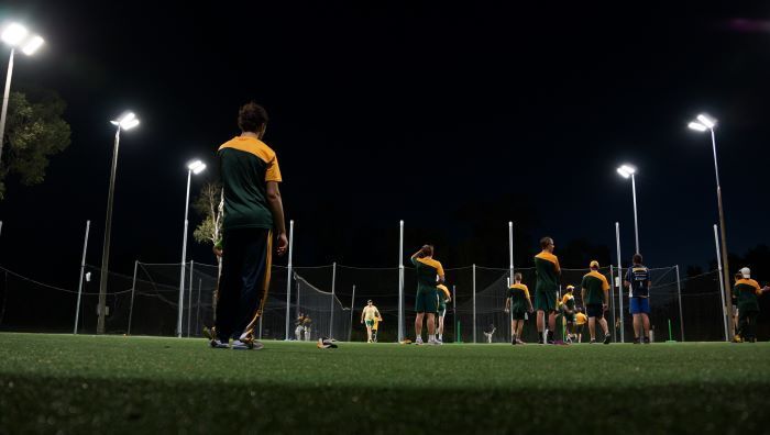 Cricket players on a field at night under bright lights; person in foreground watches the game.