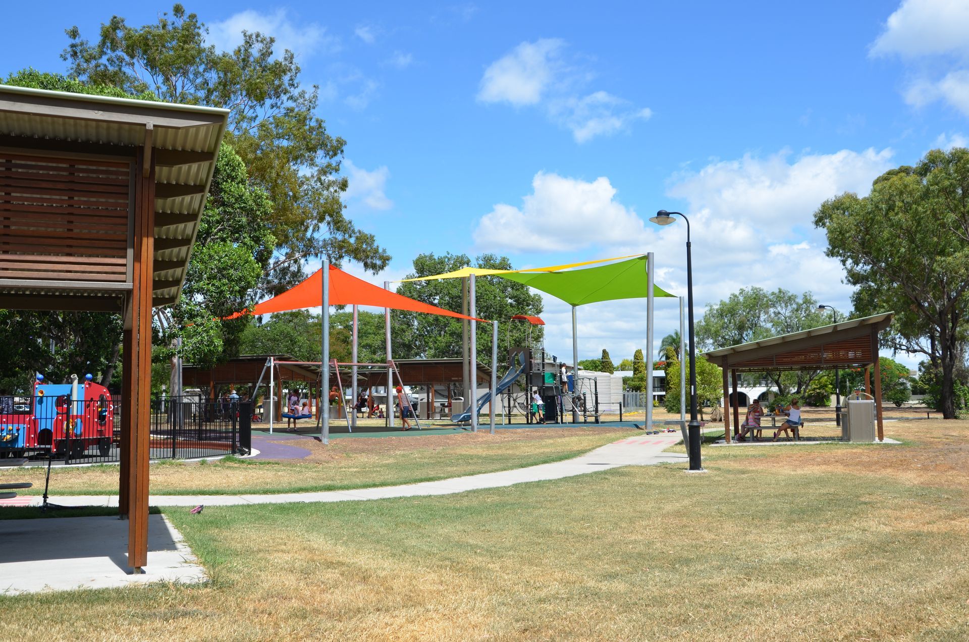 Park with playground, colorful shade sails, grassy area, picnic shelters, and blue sky.