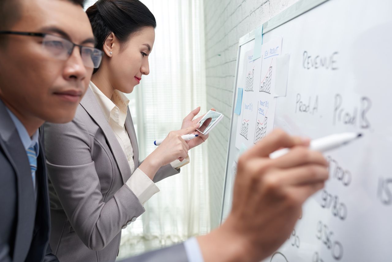Businesspeople brainstorming on a whiteboard, in an office. One writes, the other holds a phone.