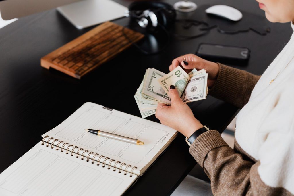 Person counting cash at a desk with a notebook, pen, phone, headphones, and wooden tray.