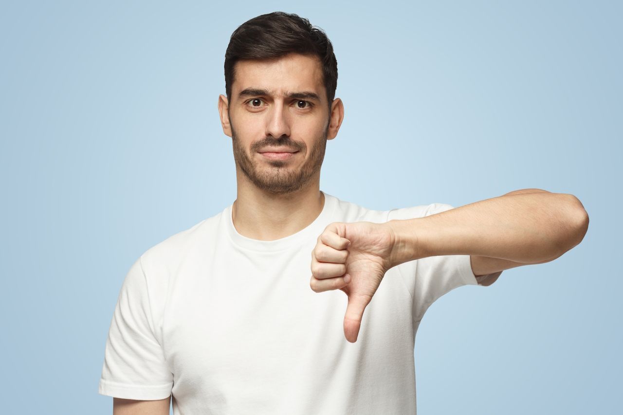Man in white t-shirt gives thumbs down gesture against a light blue background.