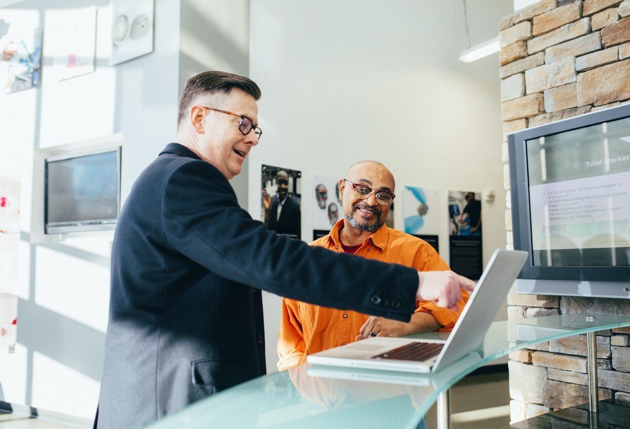 Man in suit points to laptop, showing data to a man in orange shirt at a reception desk.