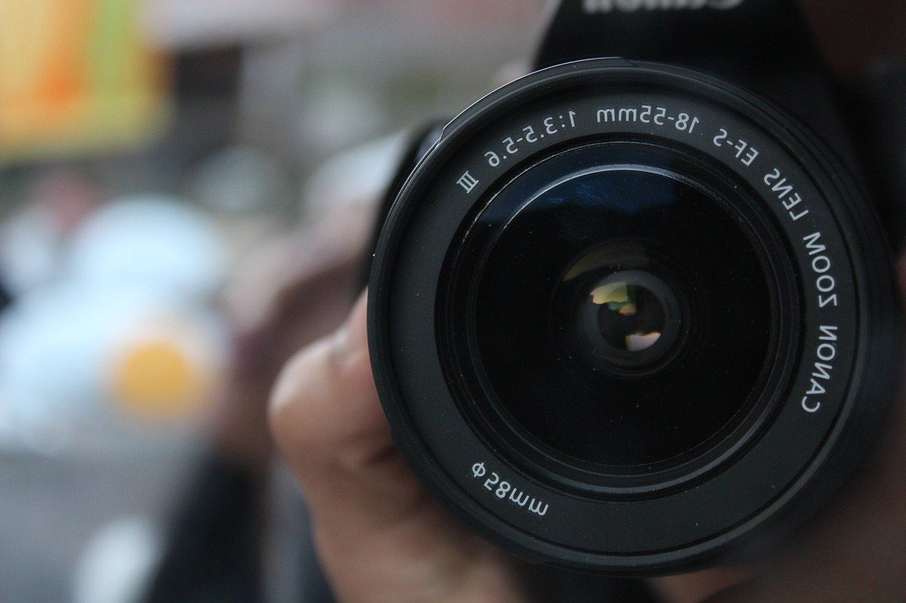 Close-up of a camera lens with text. Person holds the camera, reflected in the lens.