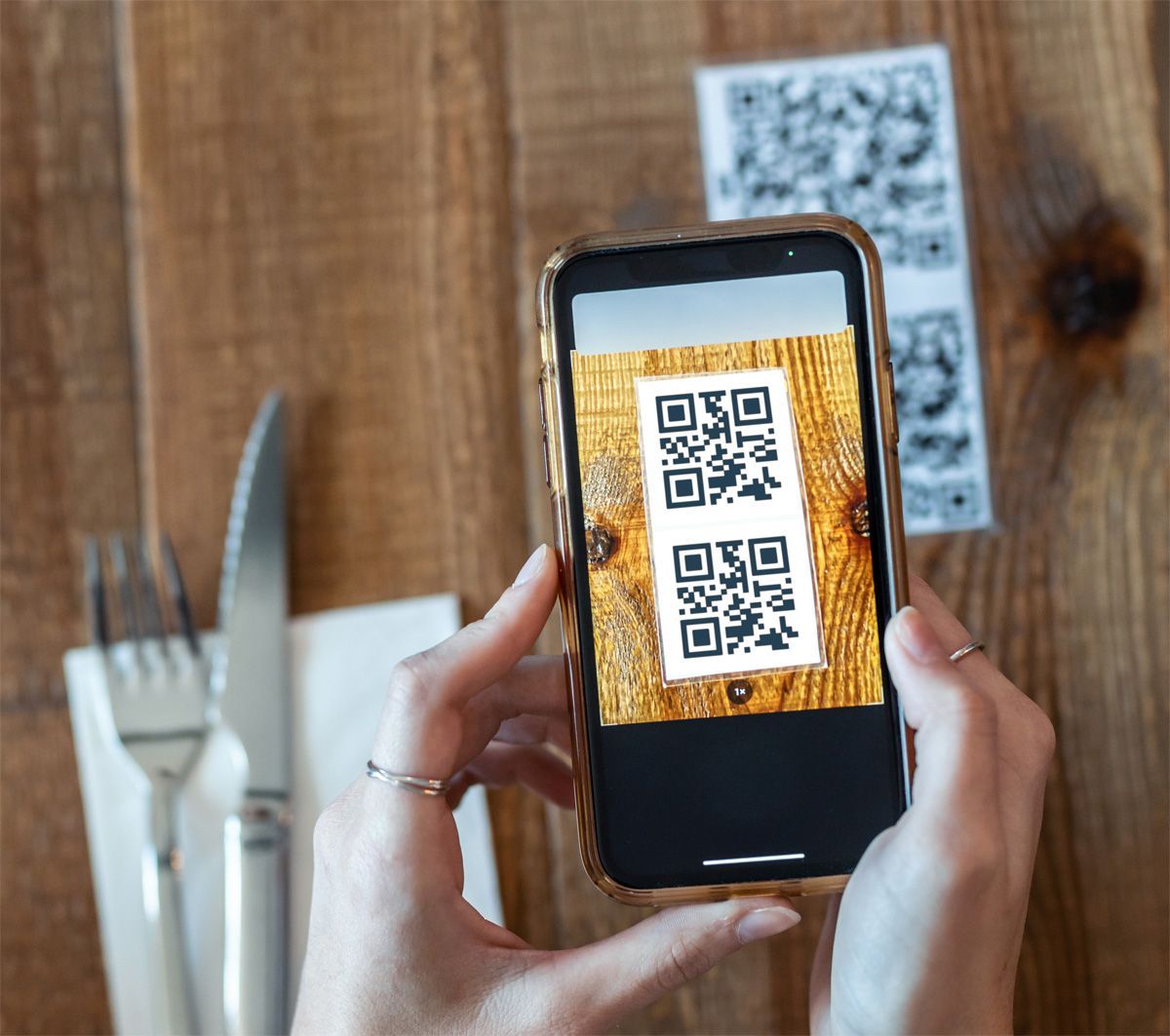 Person holding phone over a paper with two QR codes on a wooden table. Fork and knife on left.