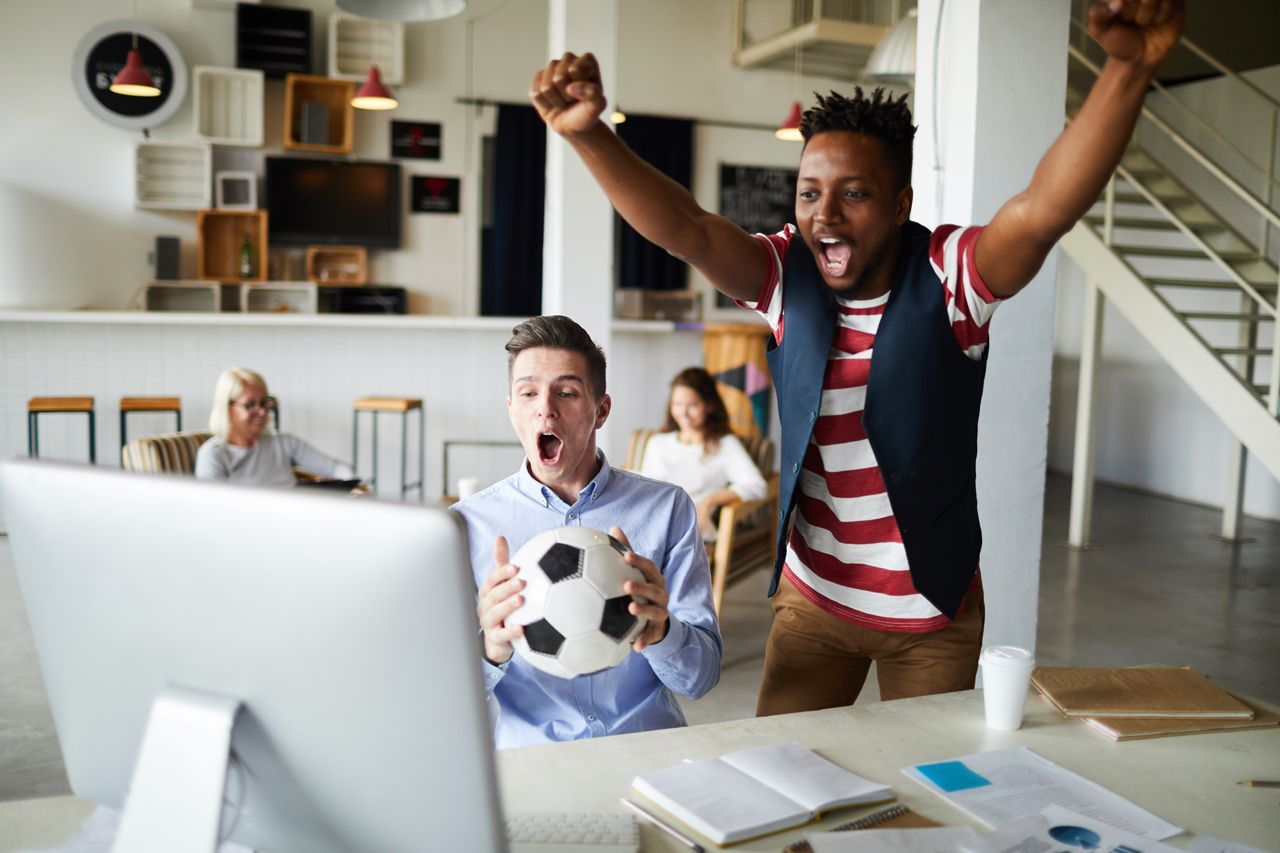 Two men celebrate with a soccer ball near a computer in an office, others watch.
