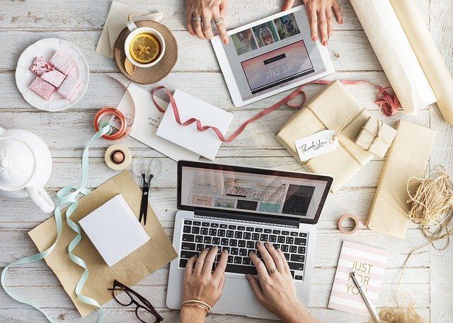 Hands typing on laptop and using tablet on a rustic, cluttered wooden desk with gifts, stationery and tea.