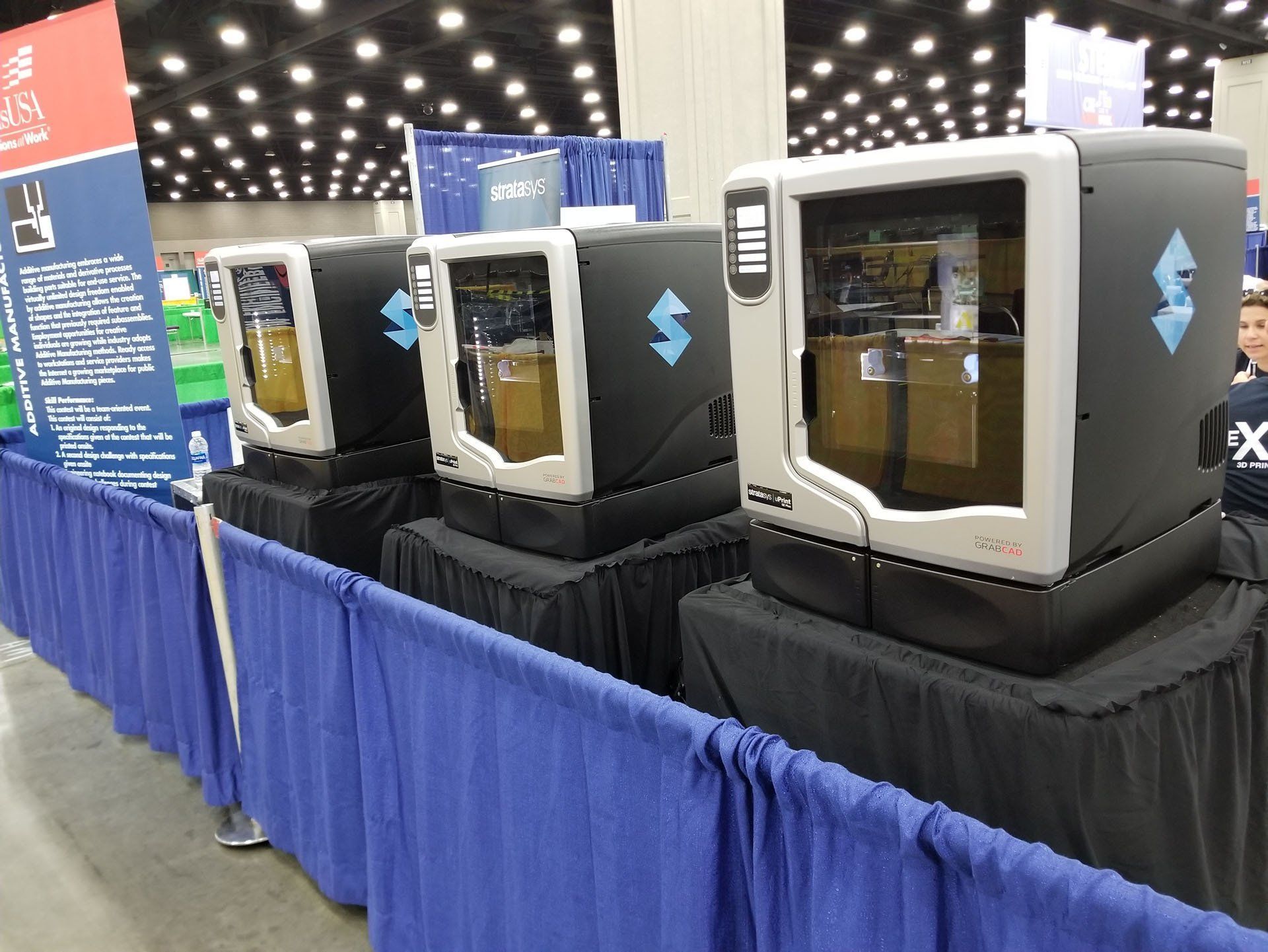 Three 3D printers with black bases and clear viewing windows at a trade show.