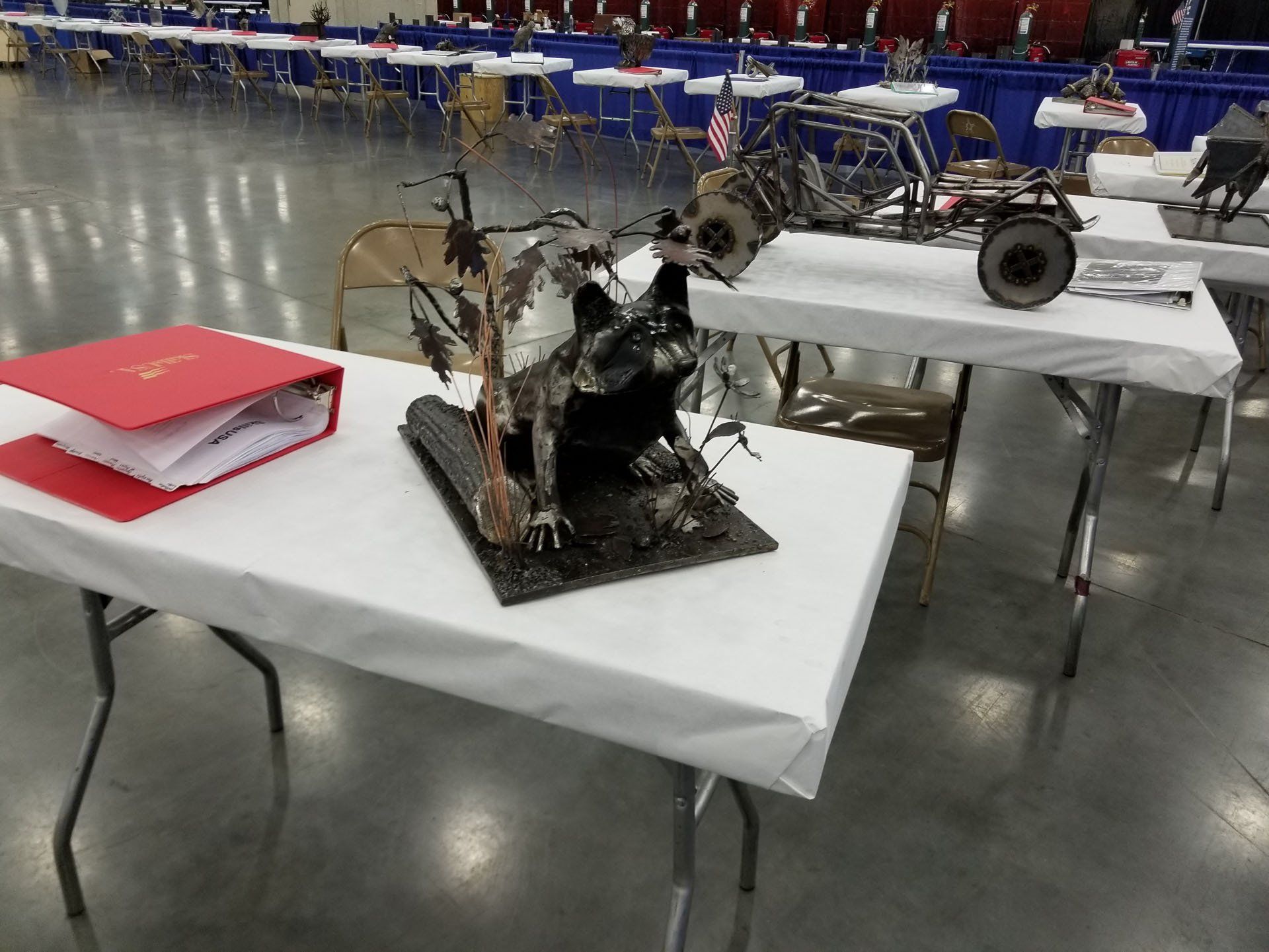 Tables displaying metal sculptures and a binder in a large hall with other tables in the background.